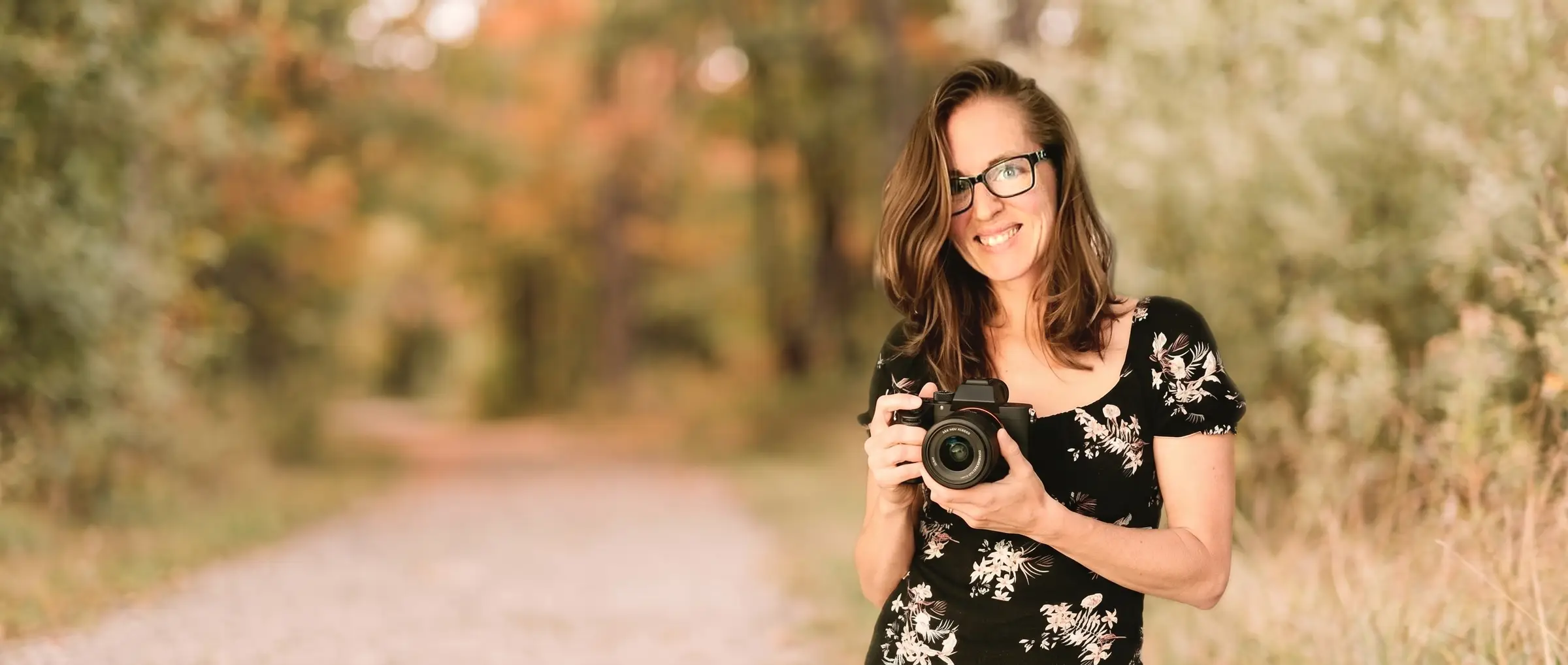 Betsy, photographer and owner of Photos by B4, holding her camera on a tree-lined park path